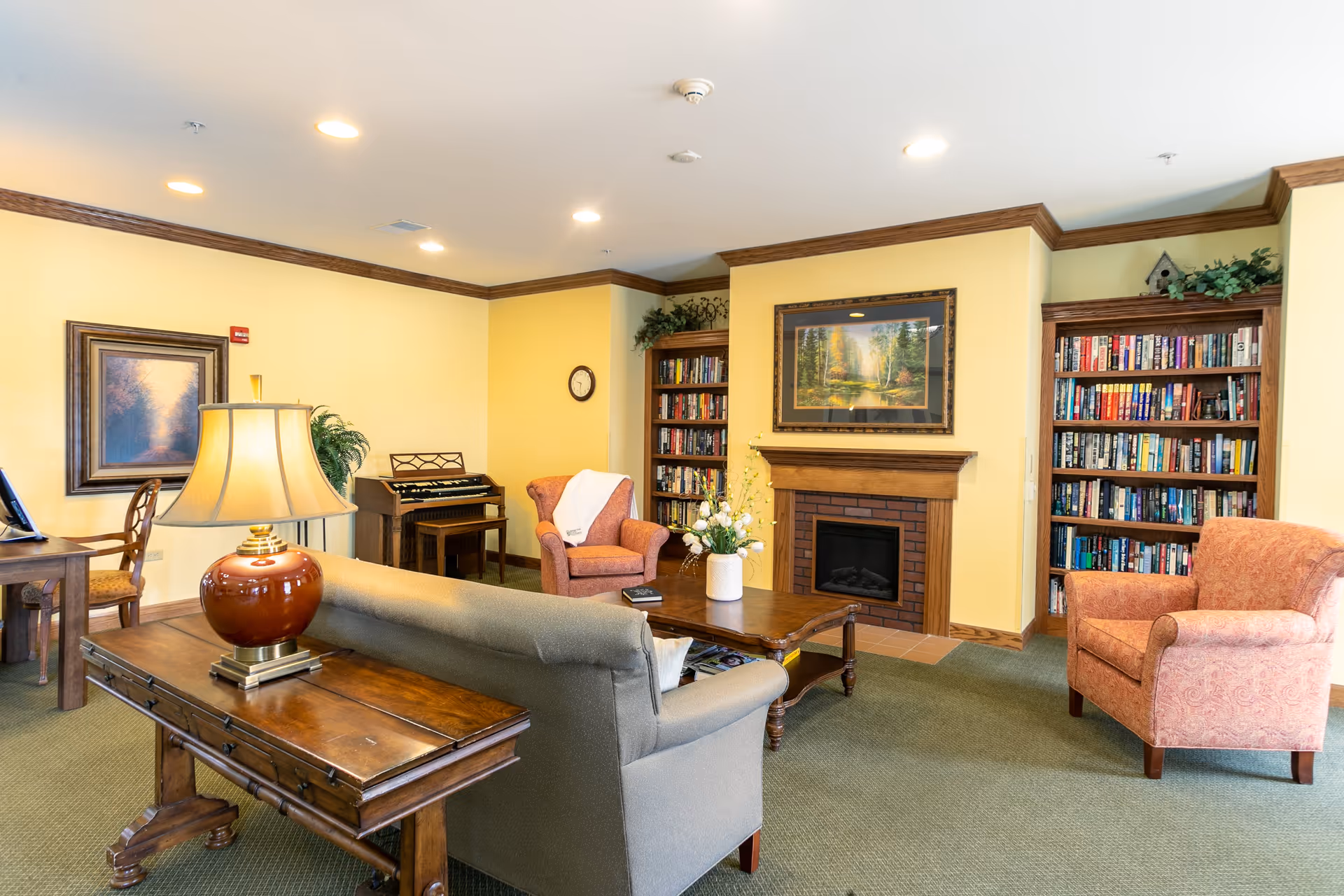 A cozy living room area in a senior living facility with a gray sofa, two upholstered armchairs, a wooden coffee table with a flower vase, and a wooden side table with a lamp. The room features a fireplace with a framed landscape painting above it, built-in bookshelves filled with books on either side of the fireplace, a small organ, and a desk with a chair. The walls are painted yellow with wooden crown molding, and the floor is carpeted in green.