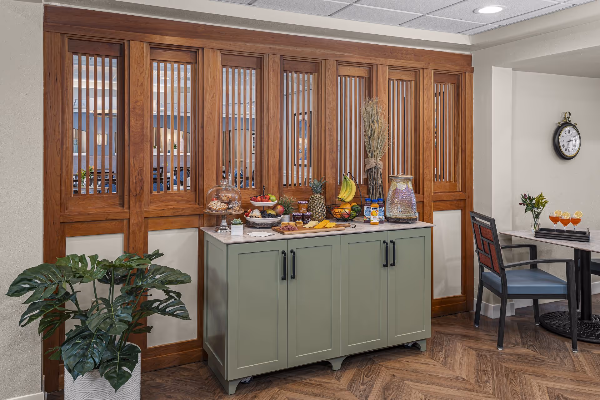 A refreshment station with a green cabinet topped with a variety of snacks including fruit, cheese, crackers, pastries under a glass dome, and beverages. Behind the station is a wooden partition with vertical slats. To the right, there is a small table with a chair and a tray holding three glasses with orange drinks garnished with lemon slices. A potted plant is placed on the floor to the left of the cabinet.