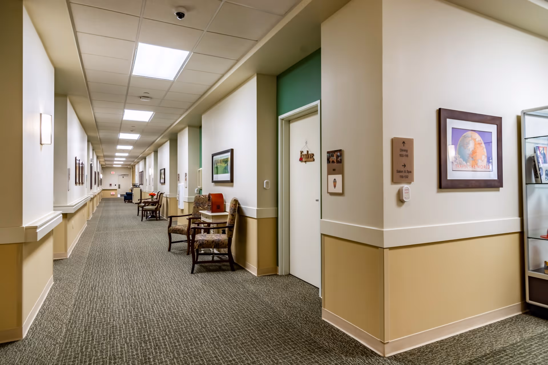 A long, well-lit hallway in a senior living facility with beige walls and carpeted floor. Several chairs and small tables are placed along the walls, with framed pictures hanging above. A door on the right side has a sign that reads 'Private'. There is a sign on the wall indicating directions to the dining area and other rooms.