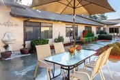 Outdoor patio area at Westgate Gardens Care Center featuring a glass-top table with six beige chairs and a large beige umbrella. The patio is adjacent to a single-story building with windows and potted plants along the wall. The ground appears wet, and there are some small bushes and landscaping around the patio.