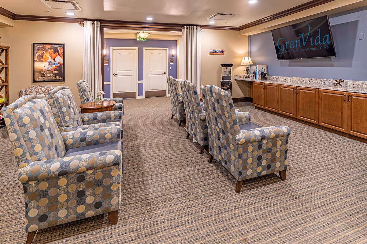 A cozy senior living common area with two rows of patterned armchairs facing a large flat-screen TV mounted on a blue accent wall. Below the TV is a long wooden cabinet with a granite countertop. The room has beige walls, carpeted floors, and a movie poster on one wall. There is a popcorn machine and a small table with a lamp near the back wall with double doors.