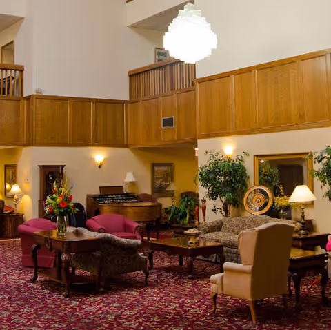 A spacious living room area with high ceilings and wooden paneling on the upper walls. The room features multiple upholstered chairs and sofas arranged around wooden coffee tables on a patterned red carpet. There are lamps, plants, and decorative items including a large plate on a stand and a floral arrangement on a table. A balcony with wooden railing overlooks the room.