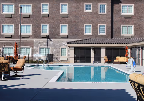 Outdoor swimming pool area at a senior living facility with lounge chairs, umbrellas, and a multi-story brick building in the background.