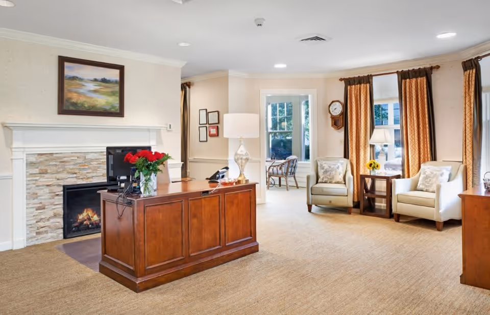 A cozy and well-lit living room area with a wooden desk in the center holding a computer monitor and a vase of red roses. Behind the desk is a fireplace with a stone surround and a landscape painting above it. To the right, there are two beige armchairs with patterned cushions, a small wooden side table with a lamp and a sunflower vase, and large windows with brown and gold curtains. The room has beige carpet and light-colored walls, creating a warm and inviting atmosphere.