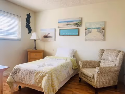 A cozy bedroom with a single bed covered in a light-colored floral bedspread. Next to the bed is a wooden nightstand with a lamp. On the wall above the bed are four framed pictures depicting beach and ocean scenes. To the right of the bed is a cushioned armchair with a patterned fabric. A window with blinds allows natural light into the room.