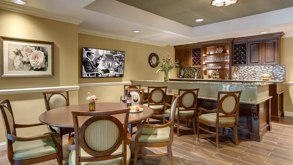 A cozy dining area in a senior living facility featuring a round wooden table with six chairs, a bottle of wine and two glasses on the table, and a bar counter with additional seating and glassware. The room has warm beige walls, a floral painting, a wall clock, and a black and white photo on the wall.
