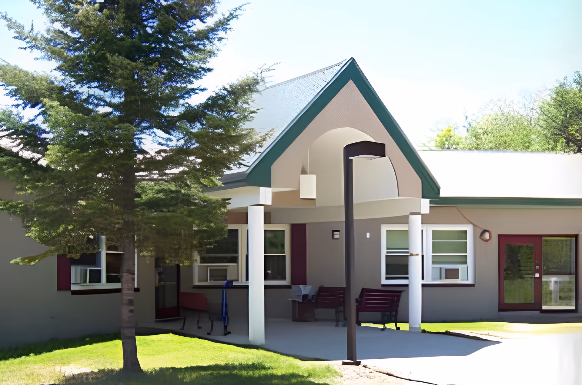 Exterior view of a single-story building with a covered entrance supported by white columns. There are benches under the entrance area, windows with air conditioning units, a tree in the foreground, and a lamp post near the entrance. The building has a gray exterior with green trim and a sloped roof.