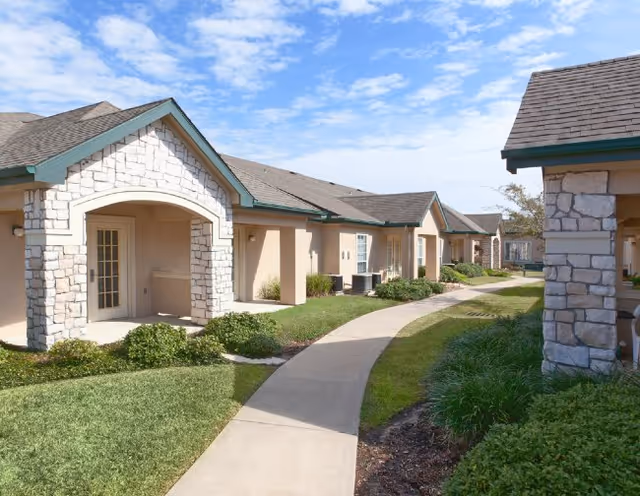 A paved walkway curves through a landscaped outdoor area with green grass, bushes, and shrubs, bordered by single-story buildings with stone and beige stucco exteriors under a partly cloudy blue sky.