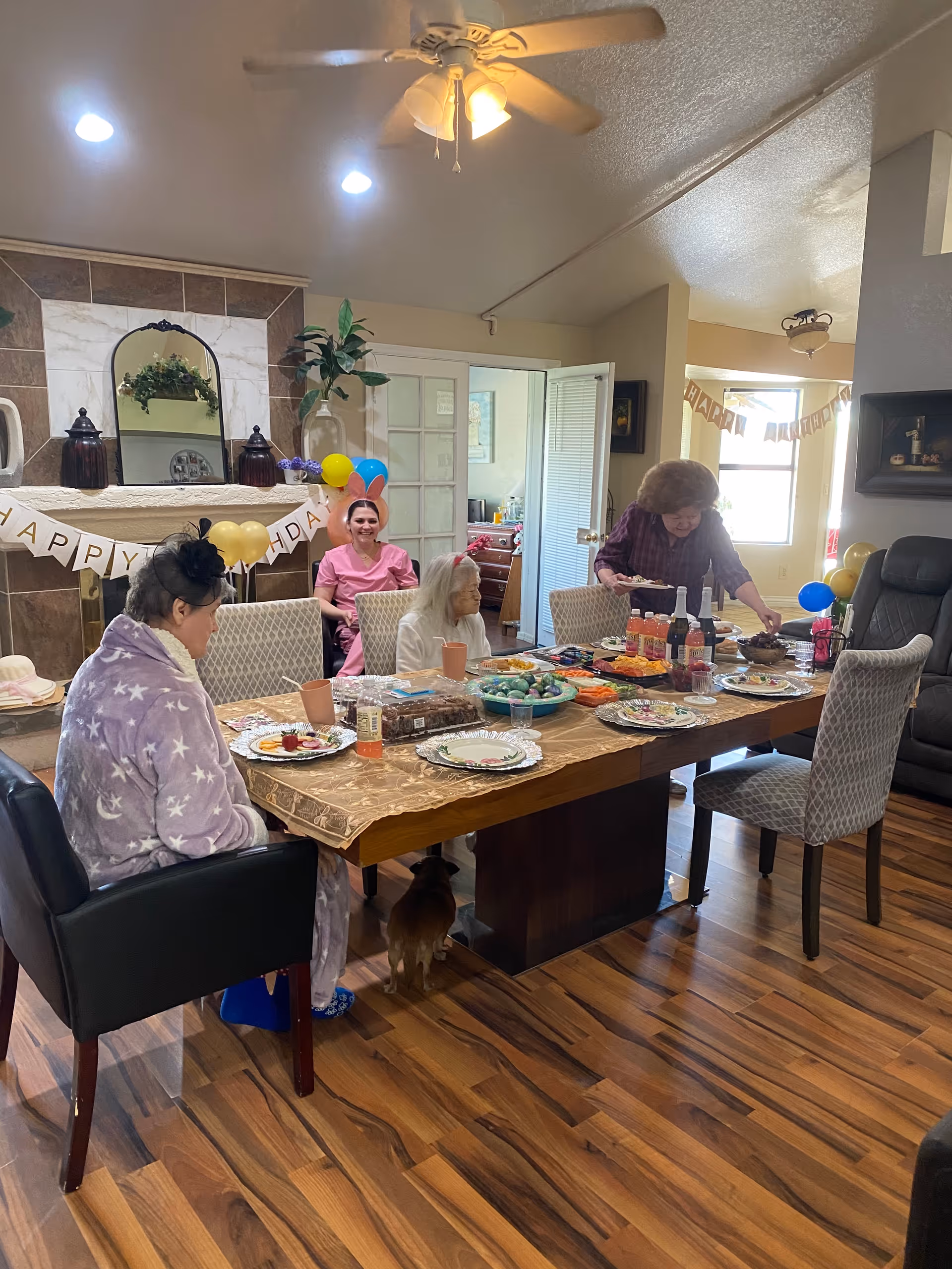 A group of elderly women and a caregiver gathered around a dining table set with plates of food and drinks in a cozy room decorated with balloons and a 'Happy Birthday' banner. The room has wooden flooring, a ceiling fan, and a fireplace with a mirror and decorative items above it.