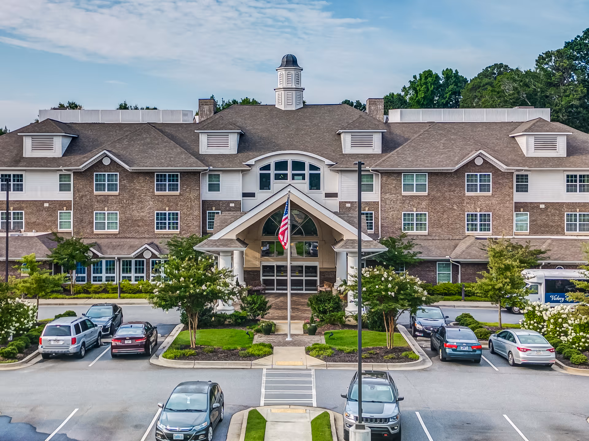 Front exterior view of Vickery Rose Retirement Resort, a large multi-story building with a peaked roof and cupola. The entrance features a covered porch with columns and an American flag in front. Several cars are parked in the parking lot, and there are landscaped trees and shrubs around the building.