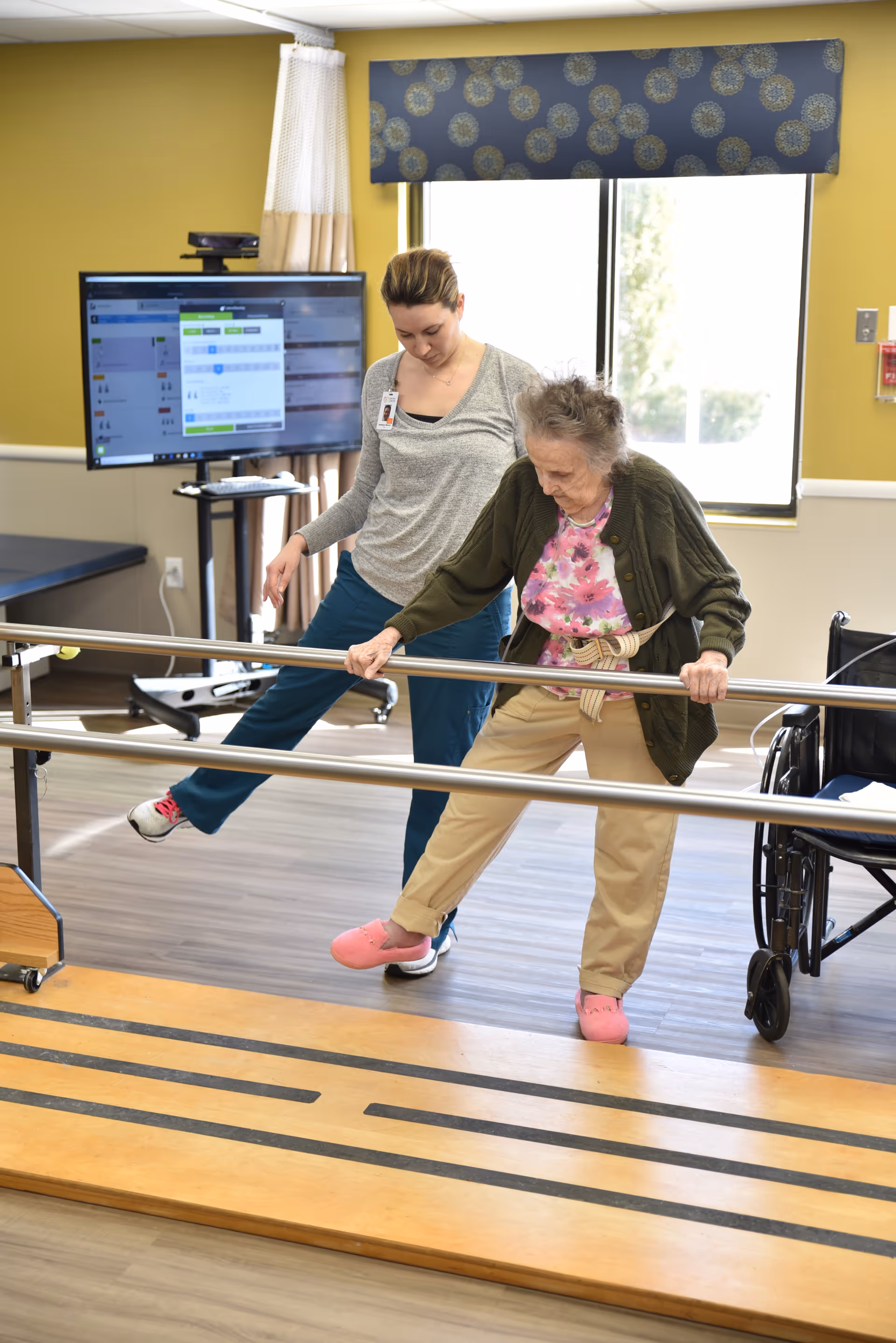 An elderly woman is performing leg exercises using parallel bars in a rehabilitation or physical therapy room, assisted by a female caregiver or therapist. The room has a large window with a blue patterned valance, a wheelchair nearby, and a monitor displaying information in the background.