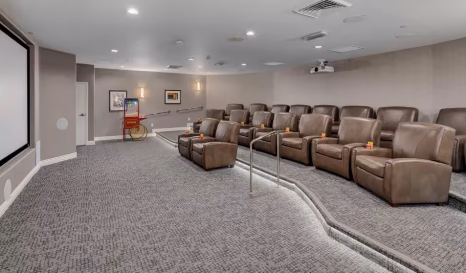 A small theater room with two rows of brown leather recliner chairs arranged on carpeted steps facing a large projection screen. There is a popcorn machine in the back corner and framed artwork on the walls. The room has recessed lighting and a ceiling-mounted projector.