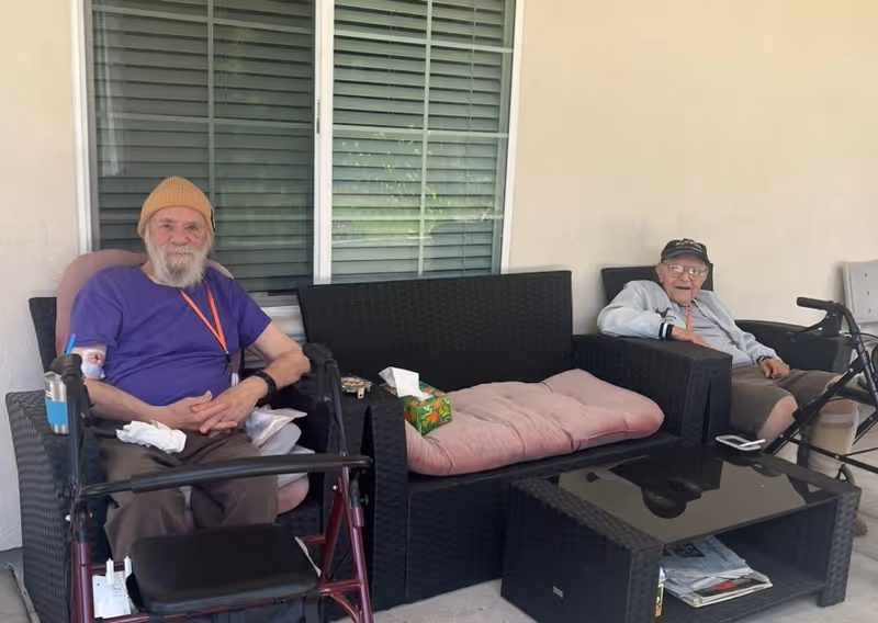 Two elderly men sit on a covered outdoor patio on wicker seating with walkers and a glass-top coffee table nearby.