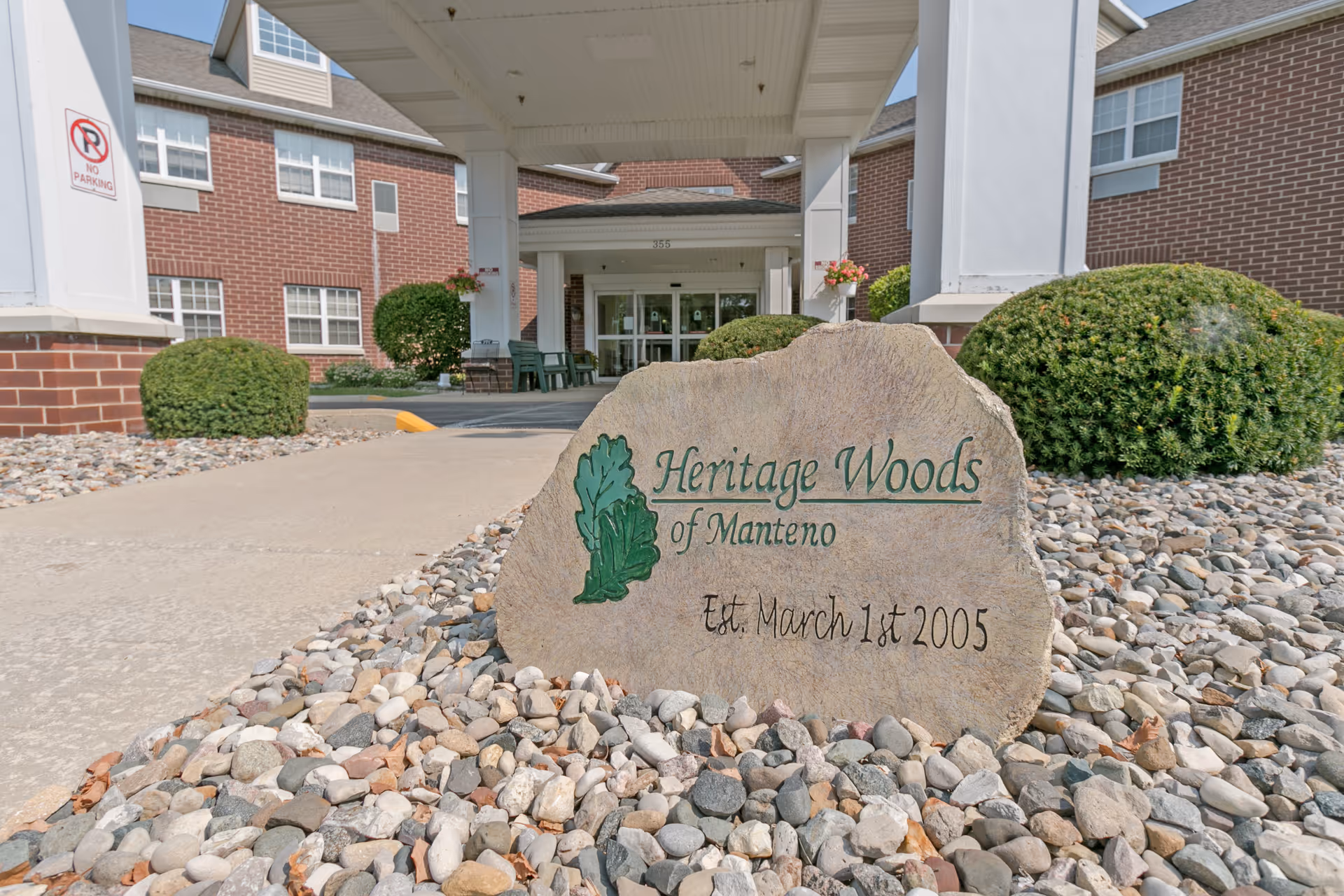 Entrance of Heritage Woods of Manteno facility with a large stone sign in the foreground that reads 'Heritage Woods of Manteno Est. March 1st 2005'. The building has a brick exterior with white pillars and a covered driveway. There are bushes and landscaping rocks around the sign and entrance.