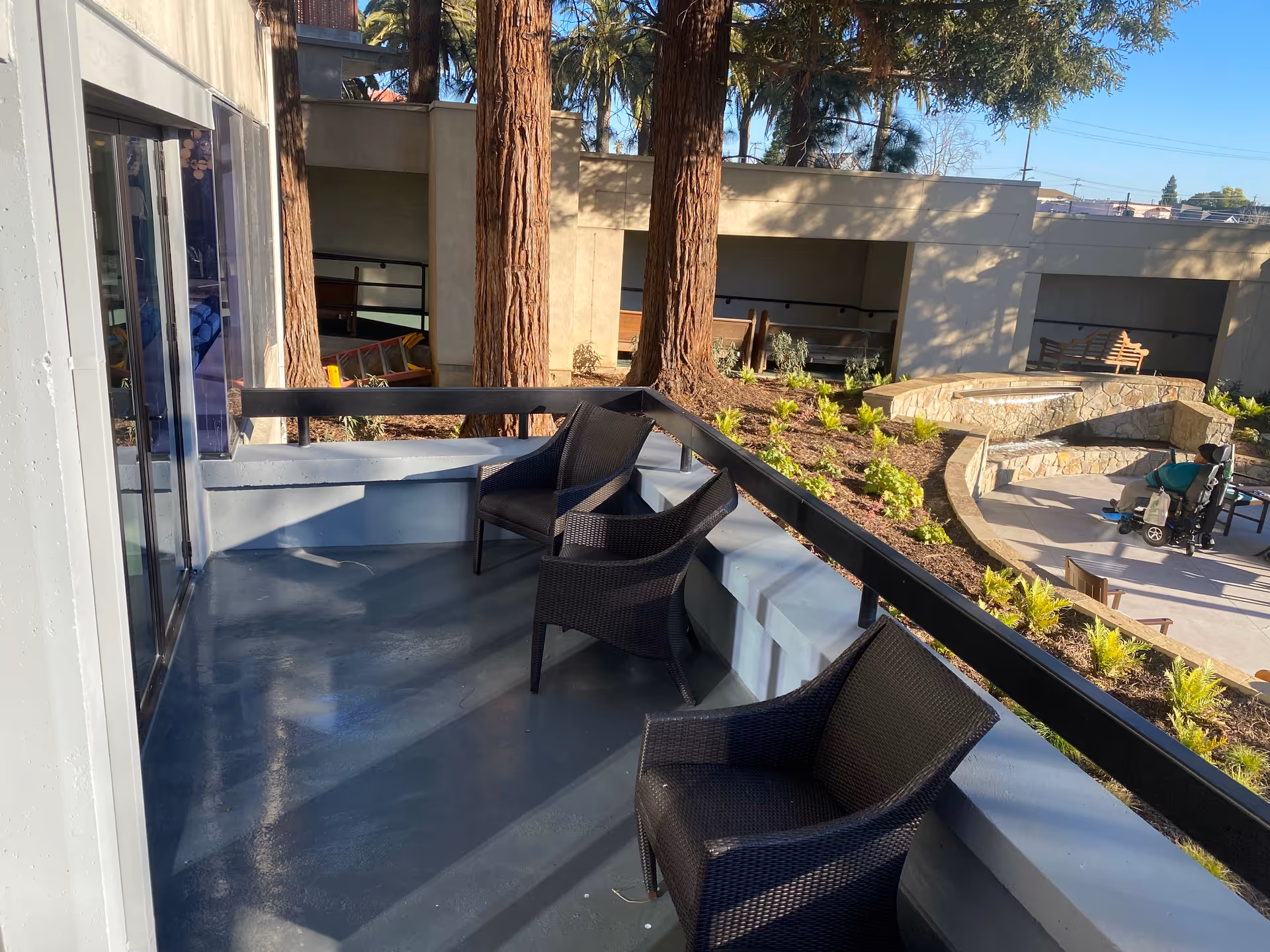 Outdoor patio area with three black wicker chairs arranged along a railing. The patio overlooks a landscaped garden with small plants, large trees, and a stone seating area. In the background, there is a person sitting on a mobility scooter near a table and chairs.