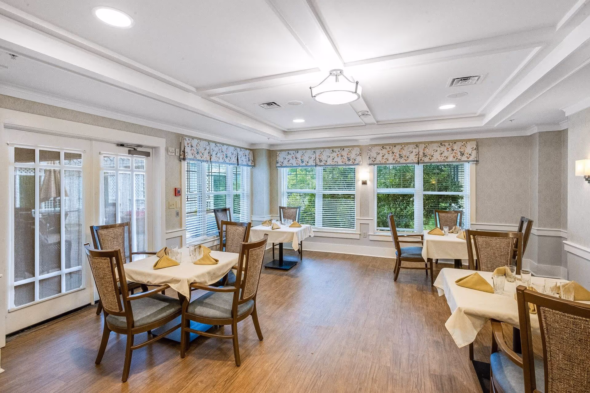A bright dining room with several tables covered in white tablecloths and set with glasses and folded napkins. The room has large windows with floral valances, wooden chairs with cushioned seats, and wood flooring. There is a ceiling light fixture and double glass doors on one side.