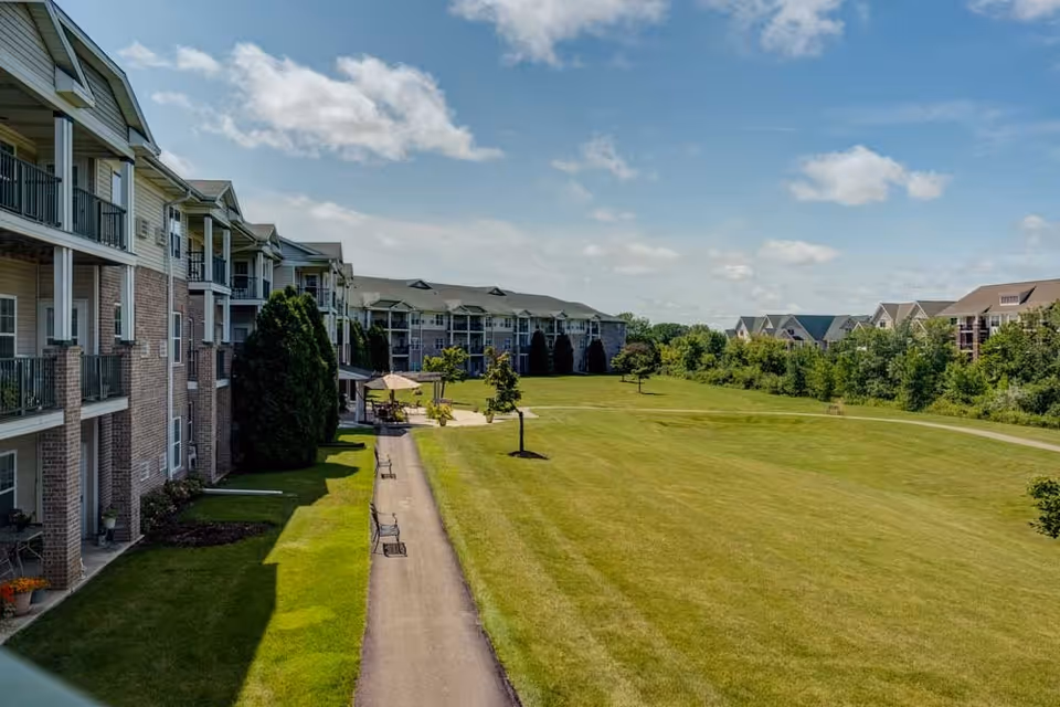 View of a senior living community with multi-story buildings featuring balconies overlooking a large, well-maintained grassy area with a paved walking path, benches, small trees, and outdoor seating with umbrellas under a partly cloudy sky.