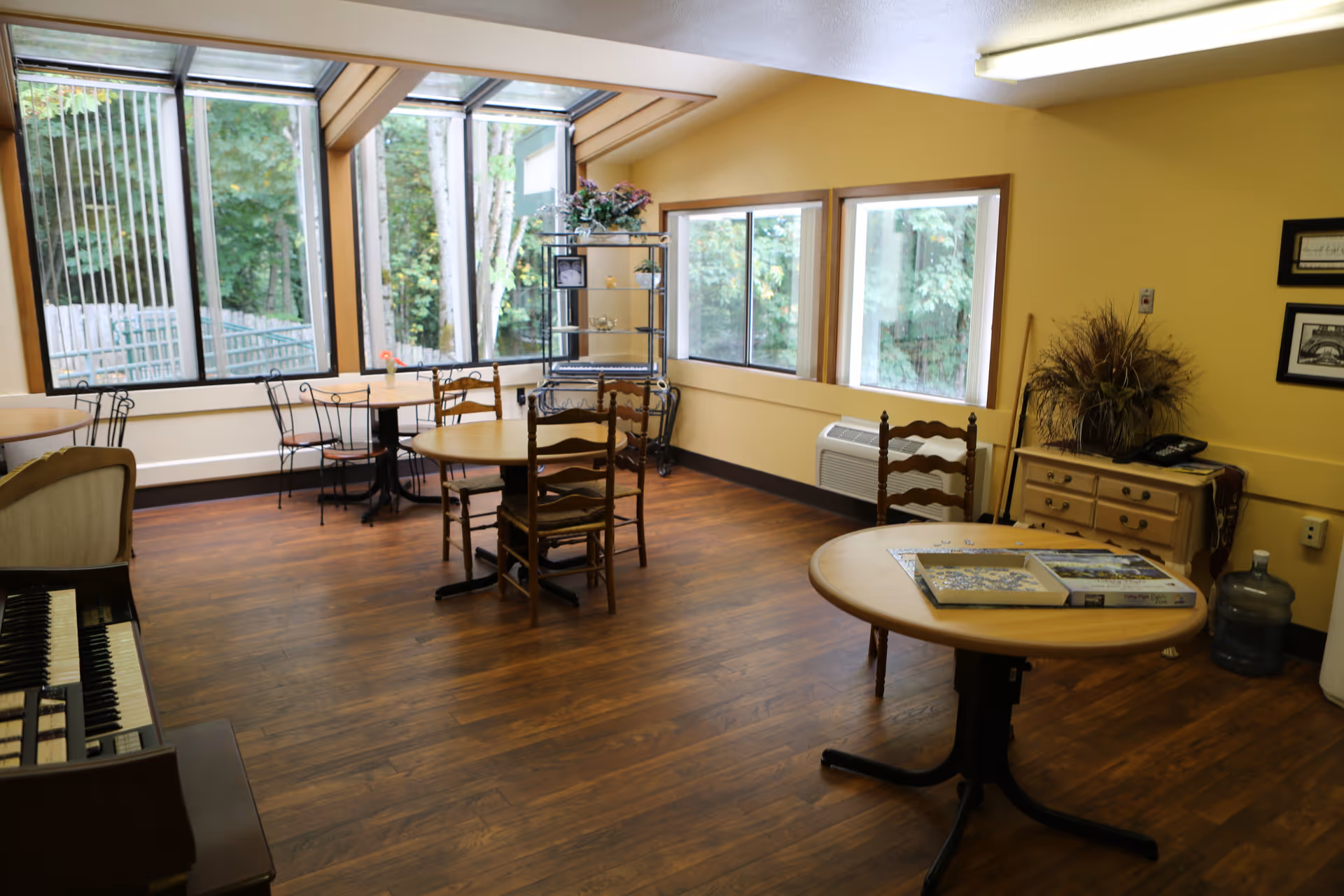A bright room with large windows showing a view of trees outside. The room has wooden floors and several round tables with chairs around them. There is a piano on the left side, a small cabinet with a phone and decorative plants on the right, and a metal shelving unit near the windows. The walls are painted yellow.