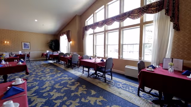 A dining room with several tables covered in red tablecloths, each set with white cups, blue napkins, and small flower vases. The room features large windows with decorative valances allowing natural light to fill the space. The floor is carpeted with a blue and gold patterned rug, and the walls have a subtle patterned wallpaper.