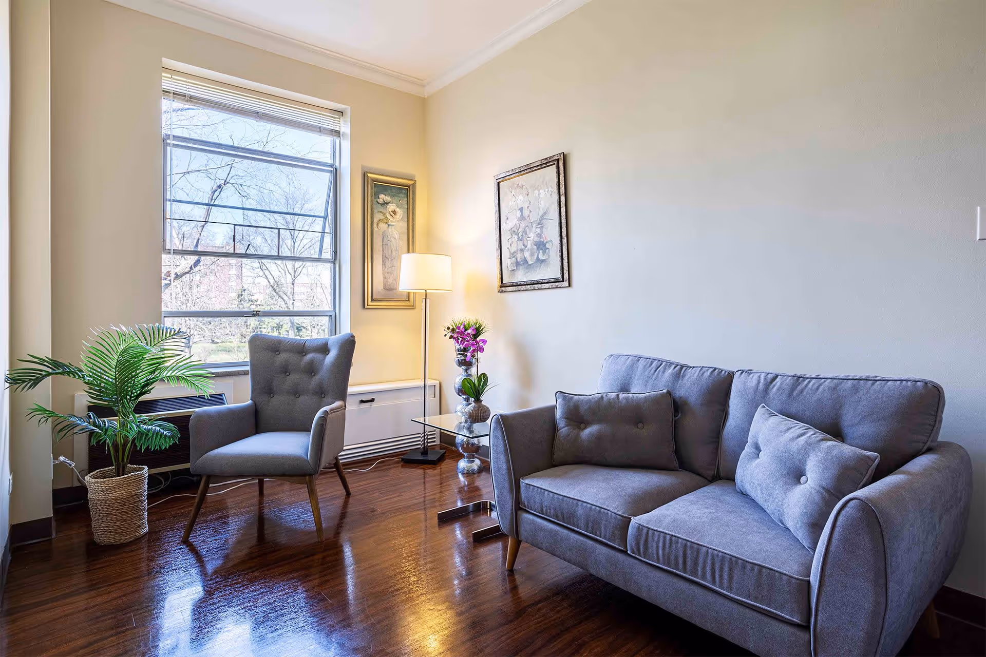A cozy living room area with a gray upholstered armchair and matching loveseat, a glass side table with a vase of purple flowers, a floor lamp, two framed floral paintings on the wall, a large window letting in natural light, and a potted plant on the wooden floor.
