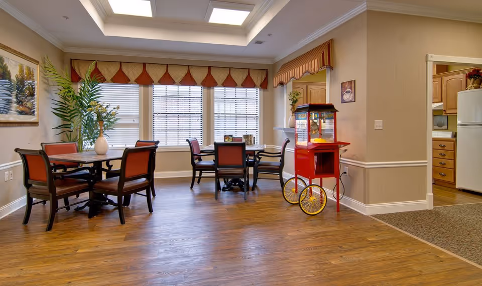 Bright dining room with tables and chairs, large windows, plants, and a red popcorn cart in a senior living facility.
