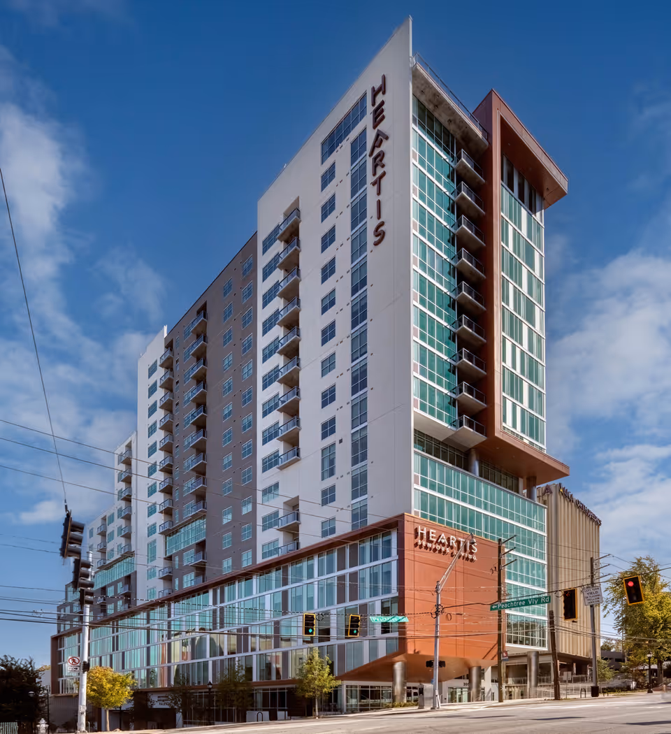 Exterior view of a modern multi-story building named Heartis Buckhead Senior Living with large glass windows and balconies, situated at a street intersection with traffic lights and street signs under a partly cloudy blue sky.