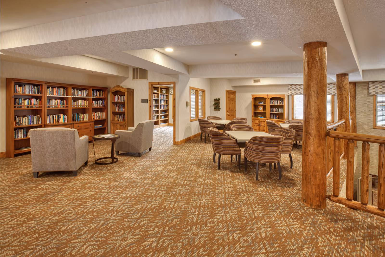 A spacious and well-lit common area in a senior living facility featuring multiple bookshelves filled with books, several comfortable armchairs, and tables with striped chairs arranged for group seating. The room has wooden pillars and railings, patterned carpet flooring, and windows with blinds allowing natural light to enter.