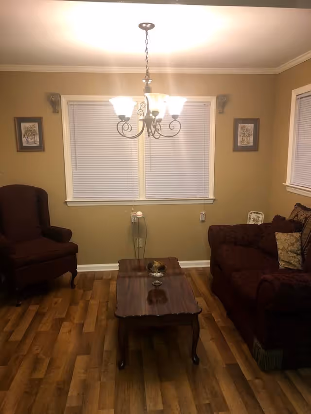 A cozy living room with wooden flooring, a dark upholstered armchair on the left, a matching dark sofa with decorative pillows on the right, a wooden coffee table in the center with a small decorative item, two windows with closed blinds on the back wall, and a chandelier with five lights hanging from the ceiling.
