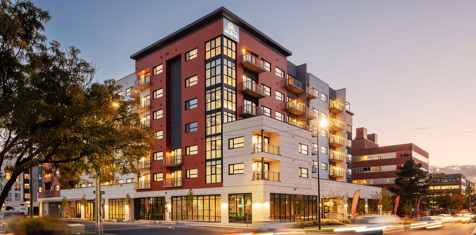 Modern multi-story residential building with balconies and illuminated street-level windows at dusk.