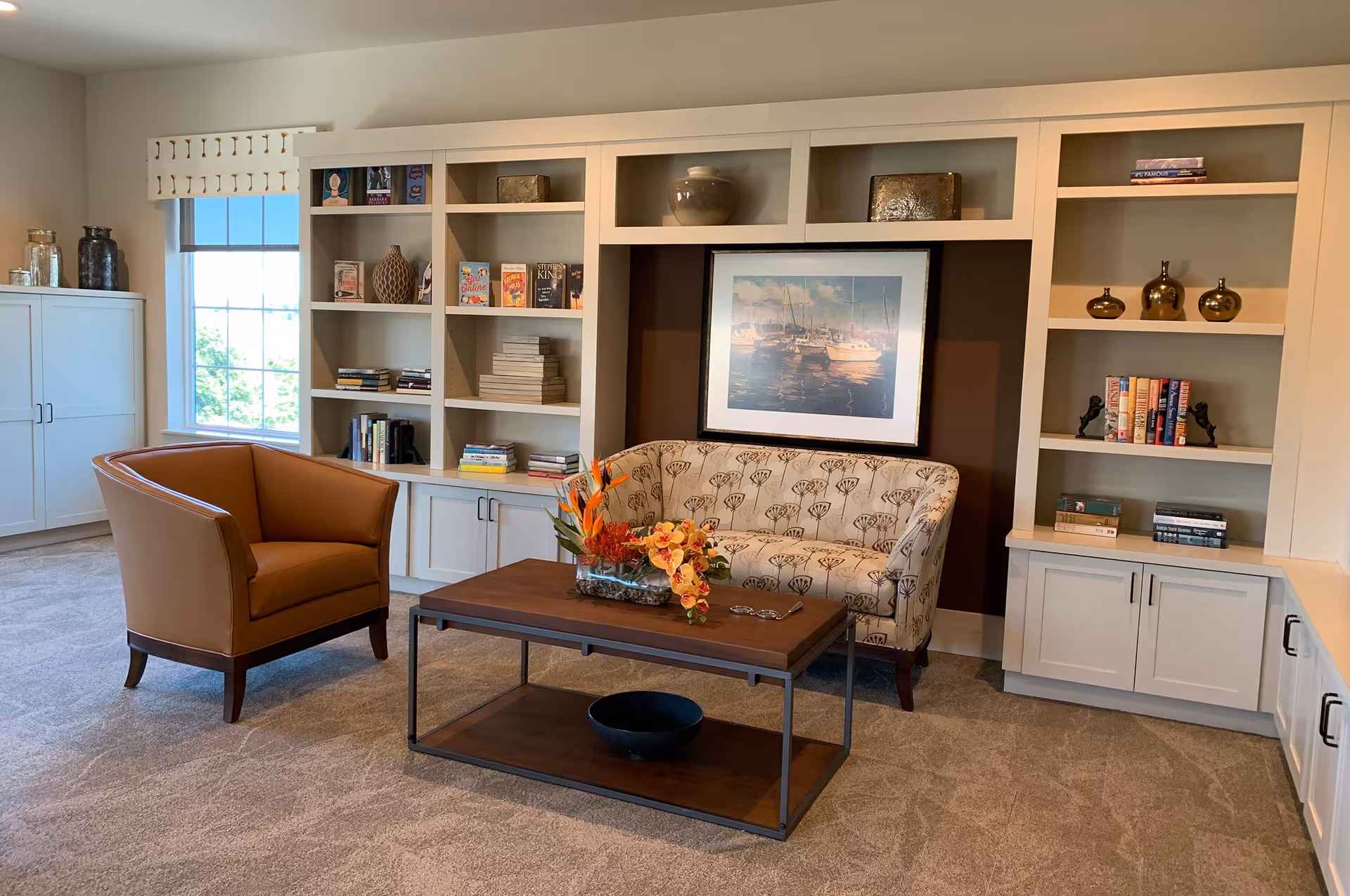 A cozy living room area with a patterned loveseat and a brown leather armchair facing a wooden coffee table adorned with a floral arrangement. Behind the loveseat is a built-in white bookshelf filled with books and decorative items, and a framed picture of boats on the water hangs on the wall. A window with a white valance lets in natural light, and the floor is carpeted in a neutral tone.