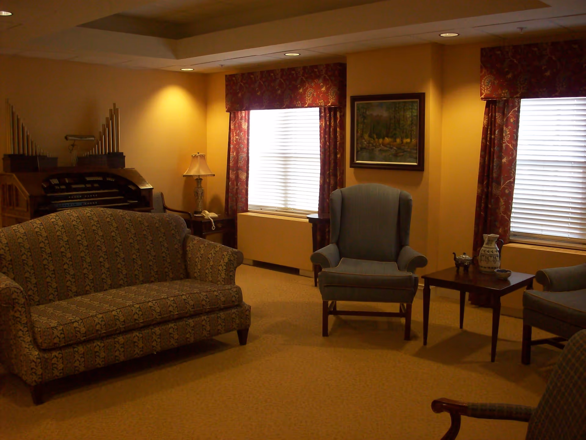 A cozy senior living room with patterned sofa and blue armchairs arranged around a wooden coffee table. The room has yellow walls, two windows with red patterned curtains, a framed painting on the wall, a table lamp, and an organ in the corner.