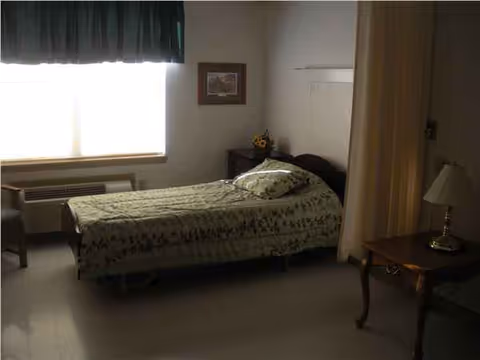 Bedroom in a senior care facility with a single bed, quilt, nightstand, and window.
