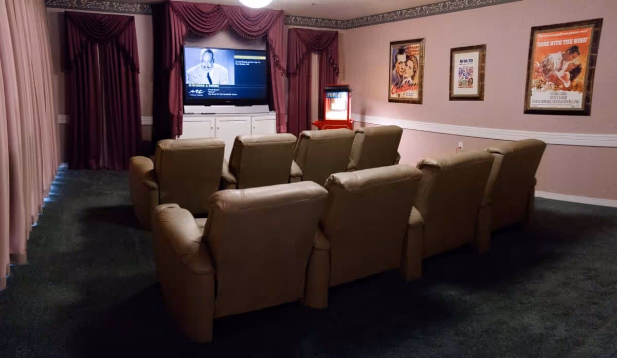 A small movie theater room with two rows of beige recliner chairs facing a flat-screen TV mounted on a white cabinet. The room has dark green carpet, pink walls with white trim, framed vintage movie posters on the wall, and burgundy curtains around the TV and windows. A red popcorn machine is visible in the corner.