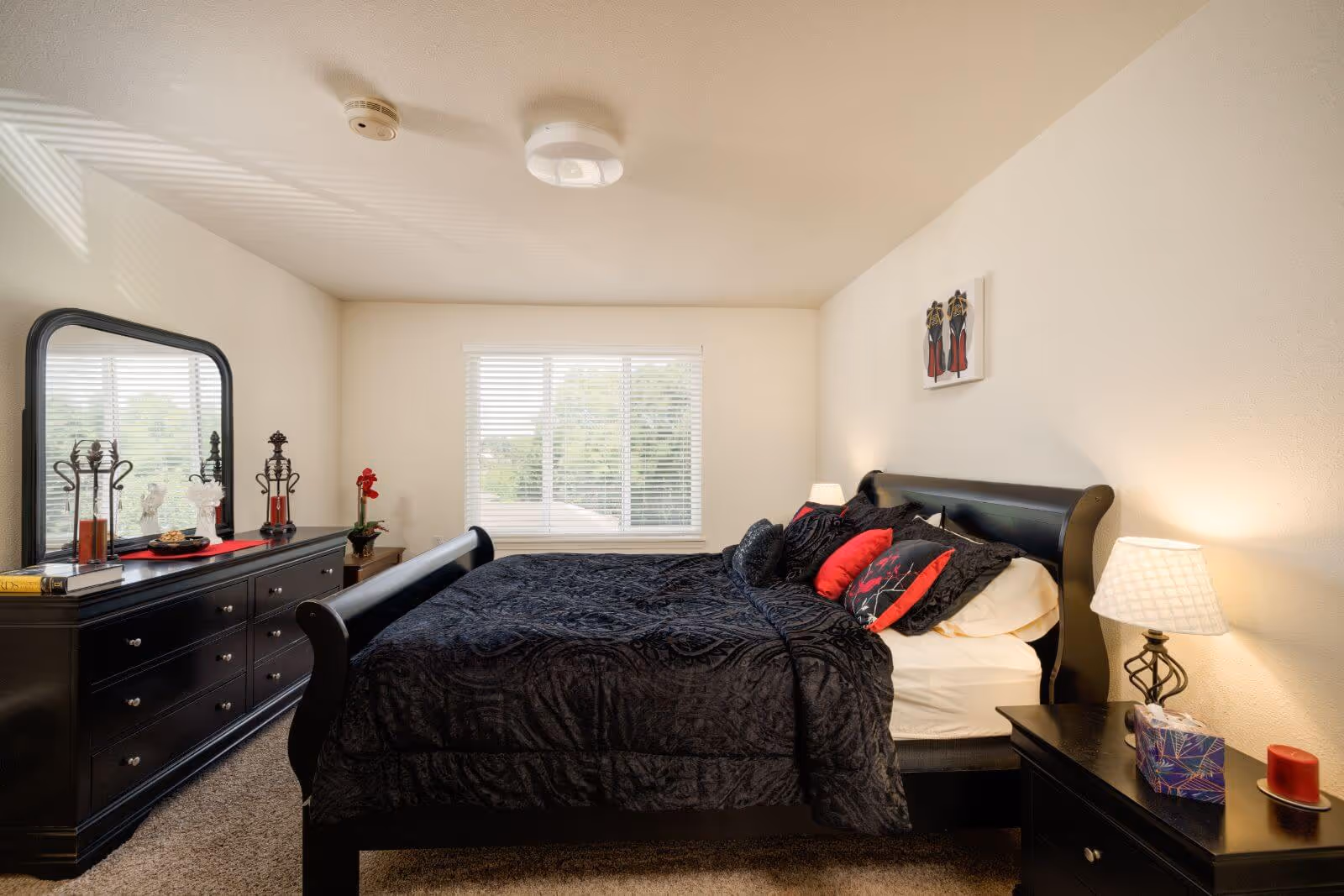Bright furnished bedroom with a black sleigh bed, matching dresser and nightstand, window with blinds and decorative accents.
