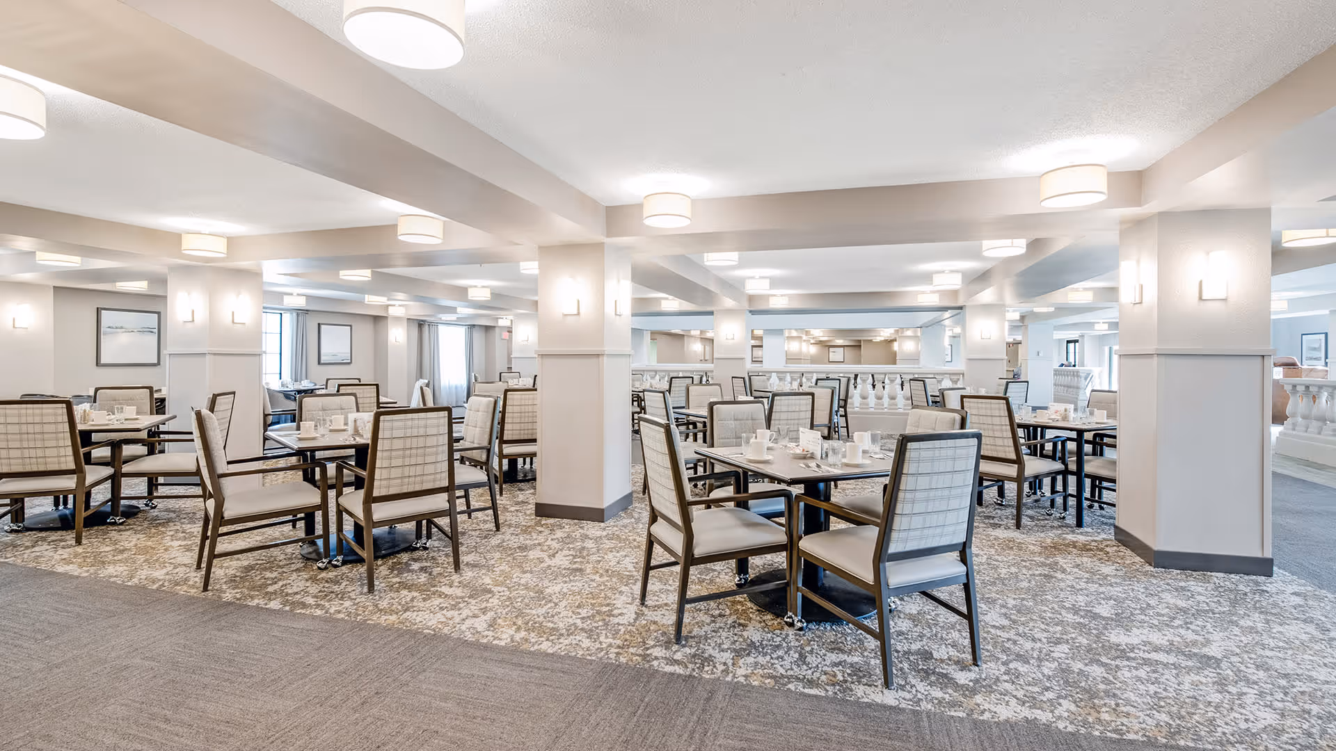 A spacious, well-lit dining room with multiple tables and chairs arranged neatly. The room features light-colored walls, carpeted floors with a patterned design, and modern ceiling lights. Each table is set with white cups and saucers, ready for use.
