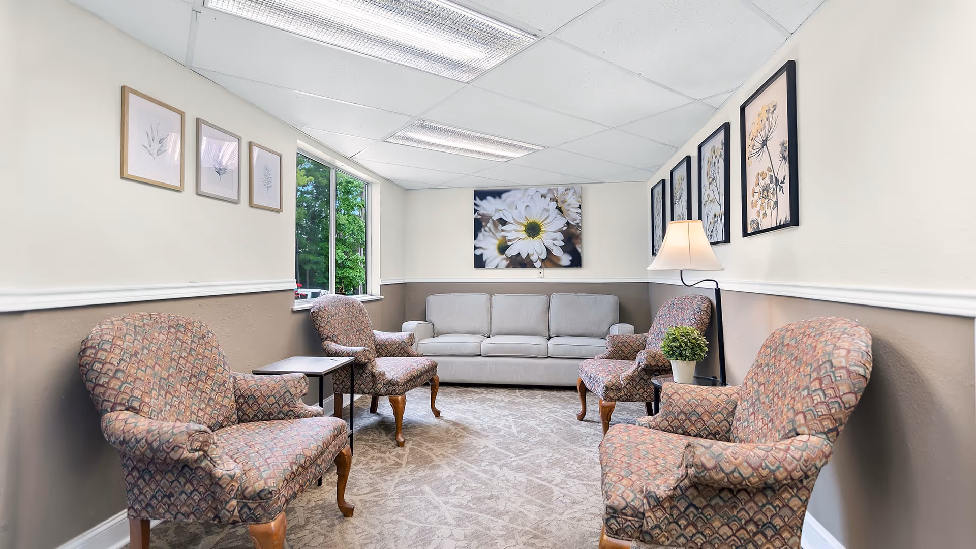A cozy sitting area in Colonial Village featuring a light gray sofa against the back wall, four patterned armchairs arranged around the room, a small side table with a potted plant, a floor lamp, and framed floral artwork on the walls. A window on the left side lets in natural light and shows greenery outside.