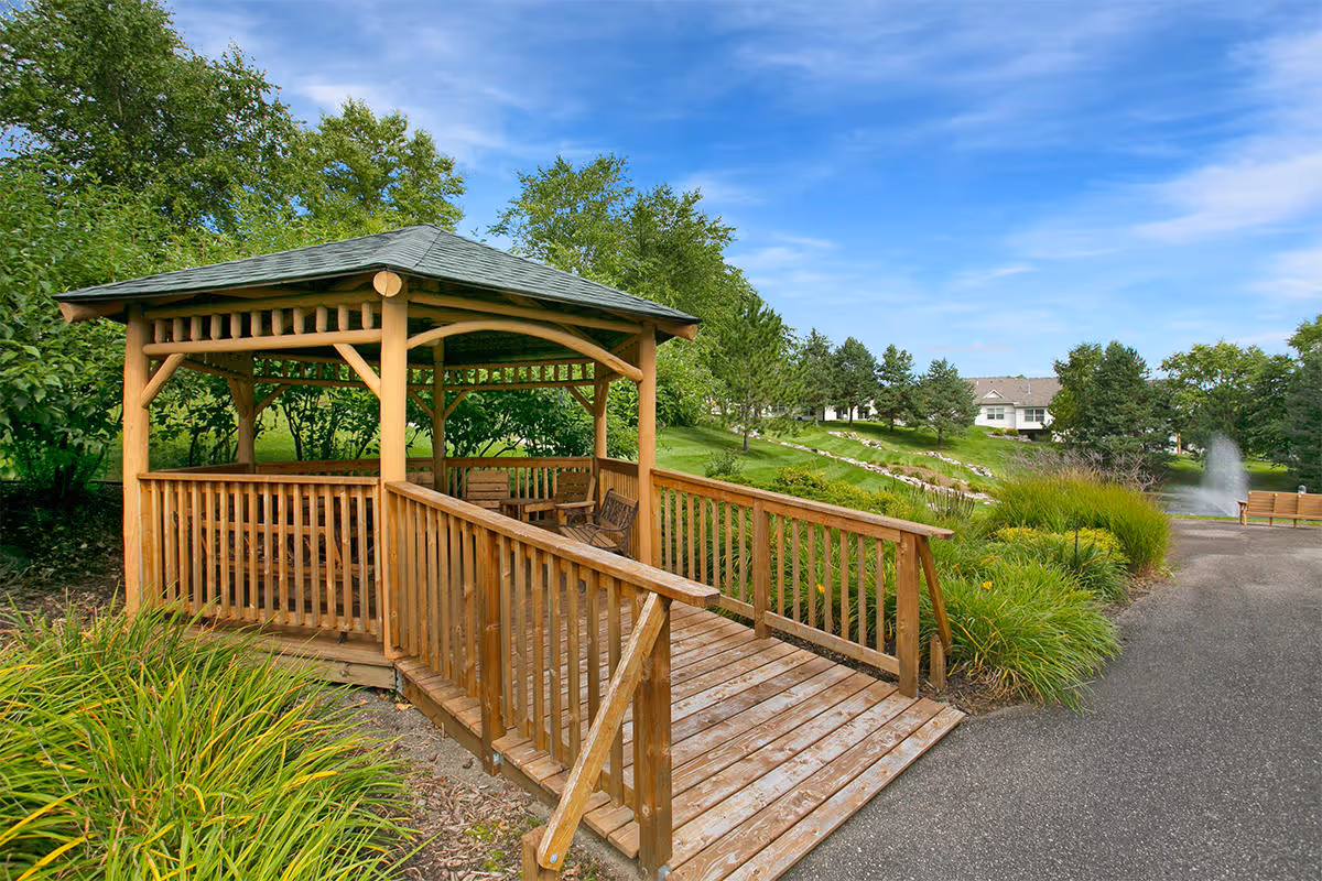 A wooden gazebo with a ramp and railing surrounded by green grass, bushes, and trees under a blue sky with some clouds. In the background, there are houses, a paved pathway, and a water fountain.