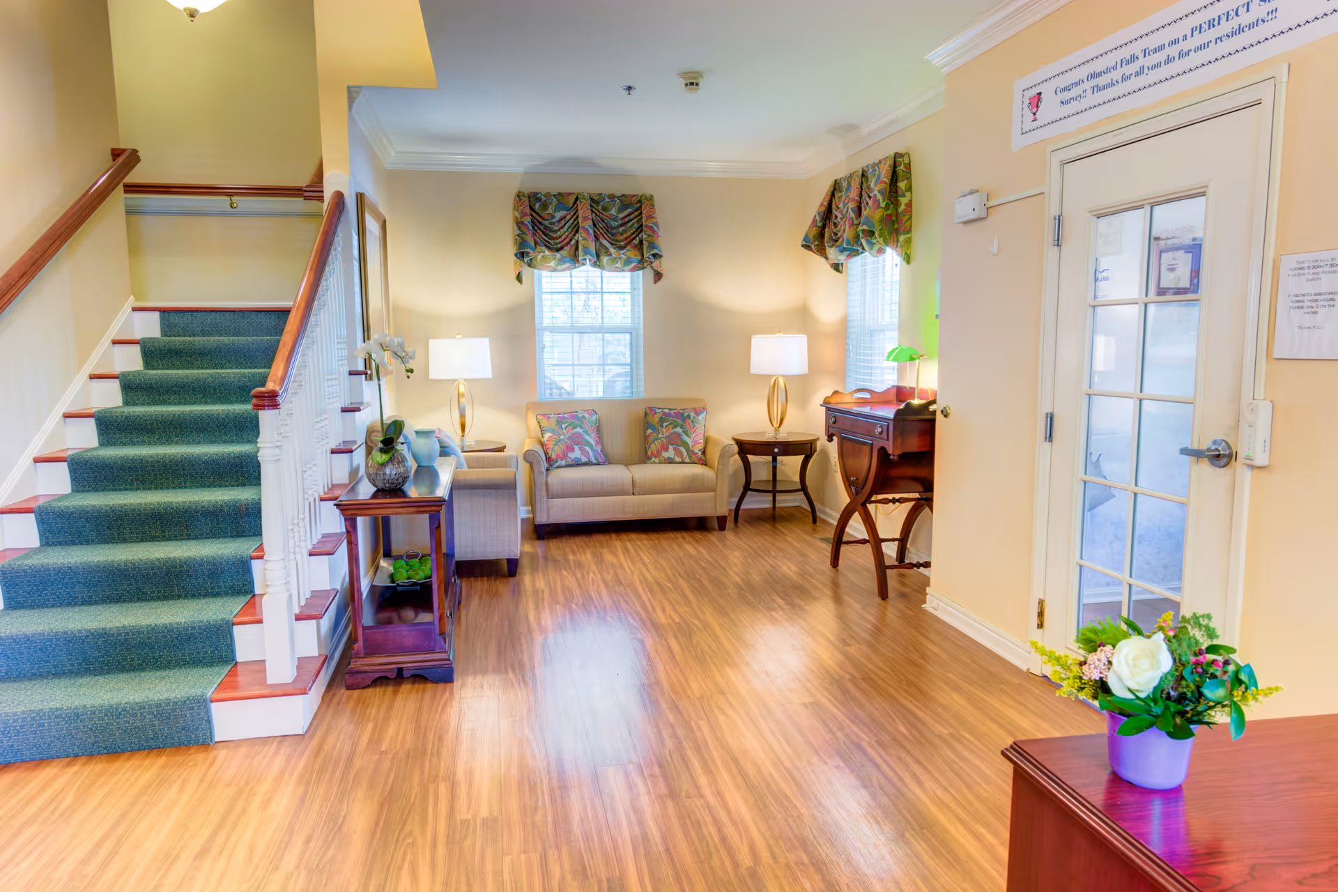 A bright and welcoming interior room with wooden flooring, a staircase with green carpet on the left, a beige loveseat with colorful cushions between two windows with patterned valances, two table lamps on side tables, a wooden desk with a green lamp, and a door with glass panes on the right. A small table with a flower arrangement is in the foreground.
