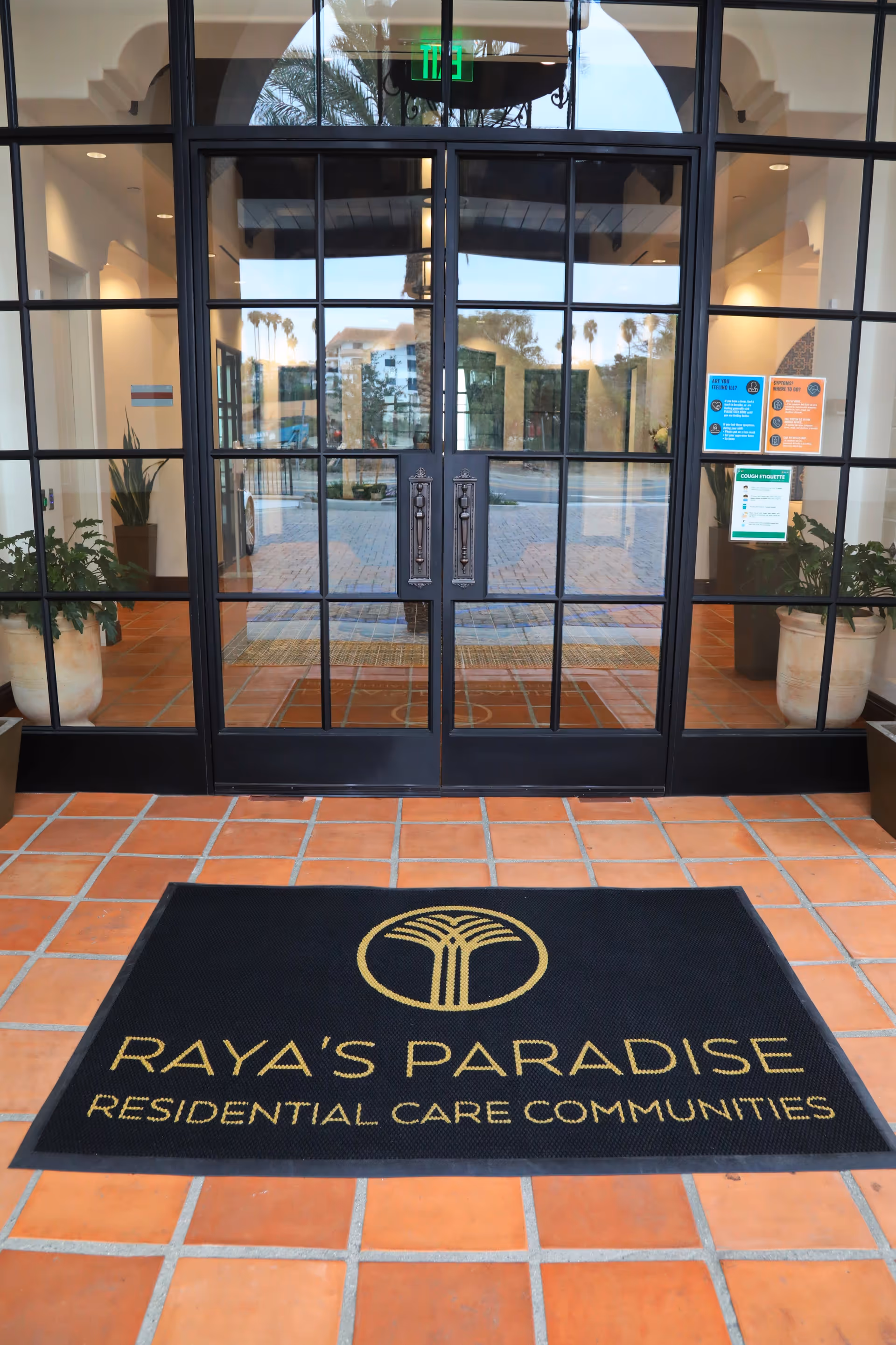 Glass double-door entrance with a black welcome mat reading "Raya's Paradise Residential Care Communities" on terracotta tile flooring.
