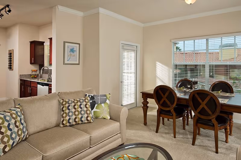 Open living room and dining area with a beige sofa and patterned pillows, a wooden dining table with chairs, and a view into the kitchen.