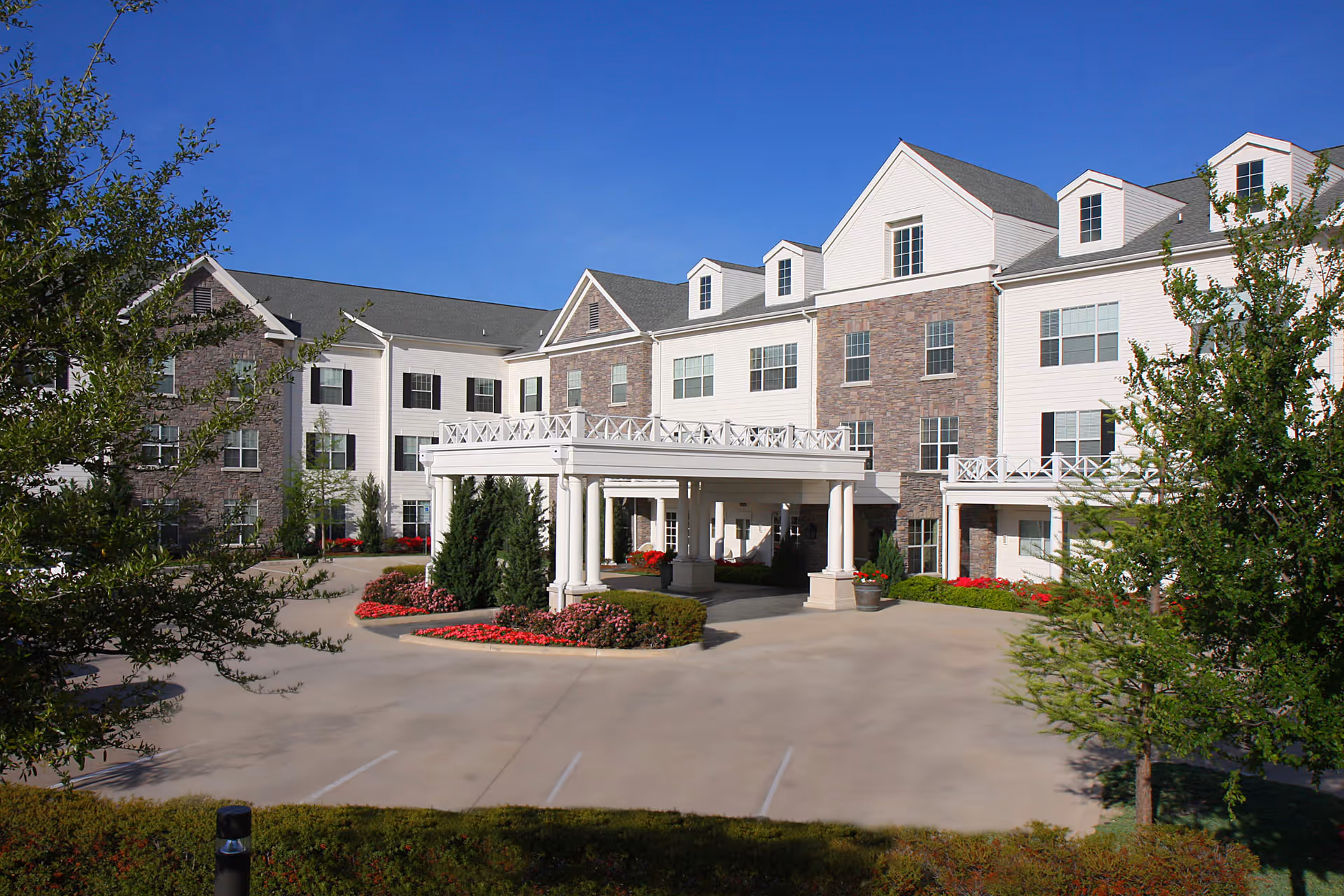 Exterior view of a multi-story senior living facility named Prestonwood Court with a covered entrance, landscaped flower beds, and trees under a clear blue sky.