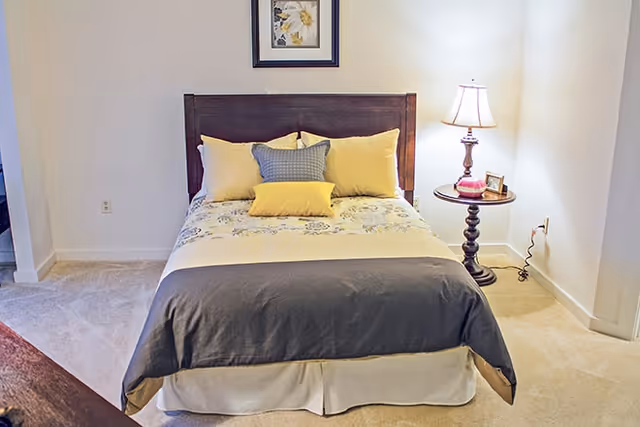 A neatly made bed with a wooden headboard, decorated with yellow and gray pillows and a floral-patterned bedspread. To the right of the bed is a round wooden side table with a lamp, a small framed photo, and a decorative item. The room has beige carpet and white walls, with a framed floral picture hanging above the bed.
