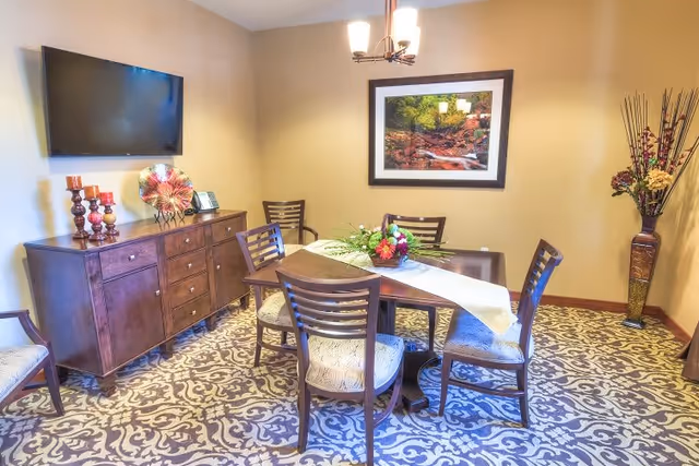 A cozy dining room with a round wooden table surrounded by five chairs with cushioned seats. The table is decorated with a floral centerpiece and a white table runner. Against the wall is a wooden sideboard with decorative items including candles and a plate. A flat-screen TV is mounted above the sideboard. The walls are painted beige, and there is a framed nature photograph hanging on the wall. The floor has a patterned carpet, and a tall vase with flowers and decorative sticks stands in the corner.