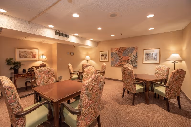 A cozy interior room with multiple wooden tables surrounded by cushioned chairs with patterned upholstery. The room is warmly lit with ceiling lights and floor lamps. The walls are decorated with framed artwork and a potted plant is placed on a small wooden desk in the corner.
