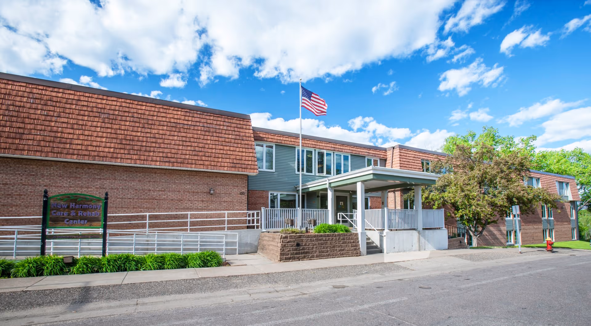 Exterior view of New Harmony Care Center, a two-story brick building with a covered entrance, an American flag on a flagpole, and a sign near the sidewalk. The sky is blue with scattered clouds and there are trees and greenery around the building.