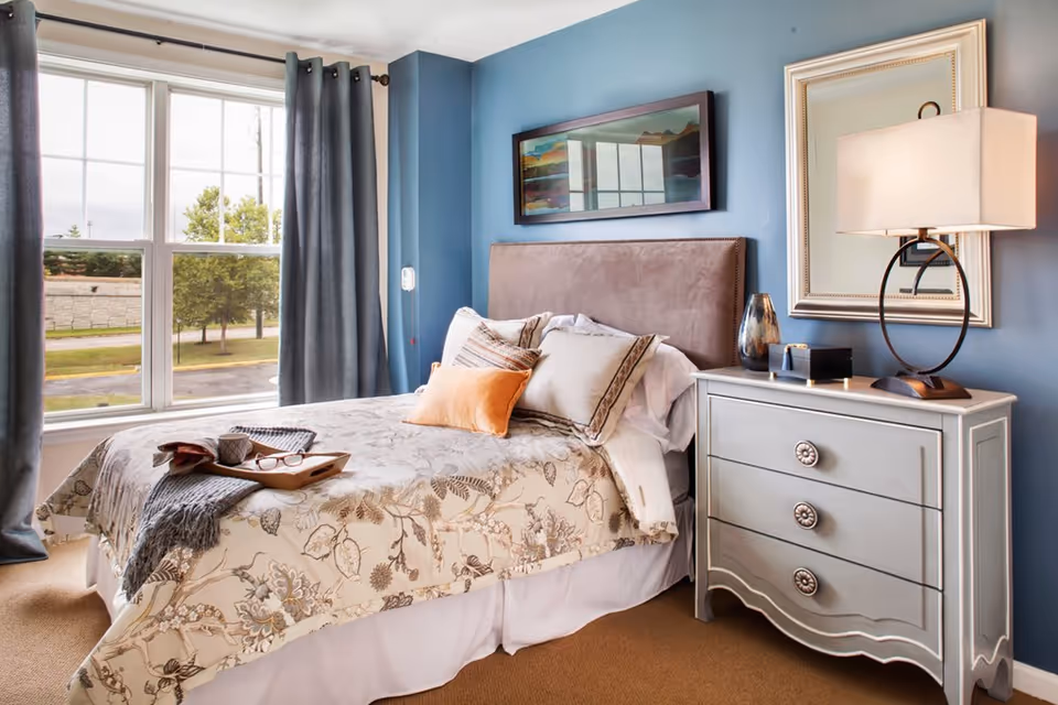 A cozy bedroom with a large window covered by gray curtains, a bed with floral patterned bedding and multiple pillows, a tray with a cup and glasses on the bed, and a gray dresser with a lamp, decorative vase, and framed mirror on the wall.
