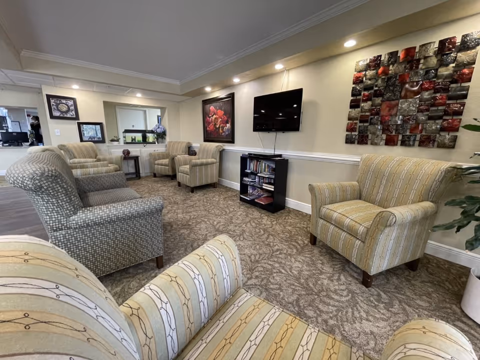 A cozy communal lounge with multiple upholstered armchairs, a wall-mounted TV, artwork, and a small bookshelf.
