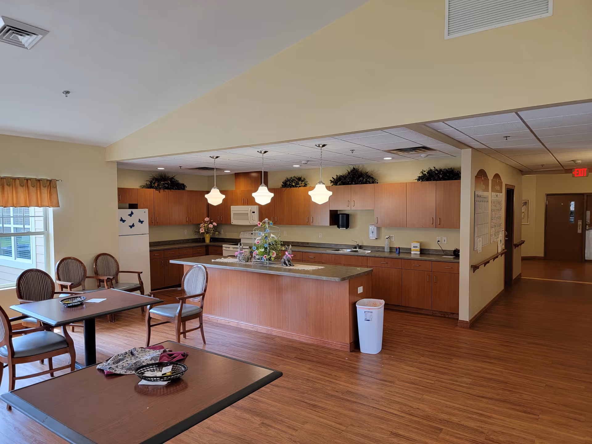 A spacious senior living facility kitchen and dining area with wooden floors, wooden cabinets, a kitchen island, hanging pendant lights, a refrigerator with butterfly magnets, and several chairs and tables near a window with a valance.