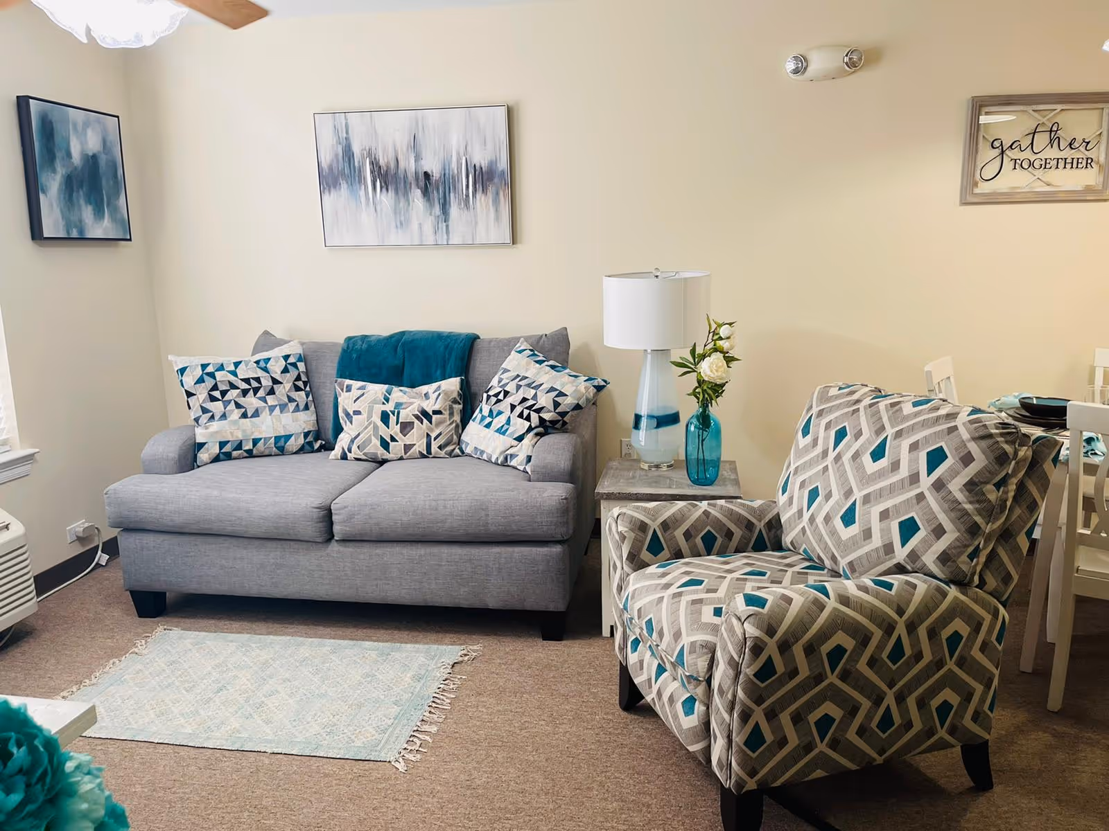 A cozy living room with a gray sofa adorned with patterned cushions and a teal throw blanket. Next to the sofa is a patterned armchair with geometric designs in gray, white, and teal. A small side table between the sofa and armchair holds a lamp and a blue vase with white flowers. The walls are decorated with abstract paintings and a framed sign that reads 'gather TOGETHER'. A light-colored rug lies on the carpeted floor in front of the sofa.