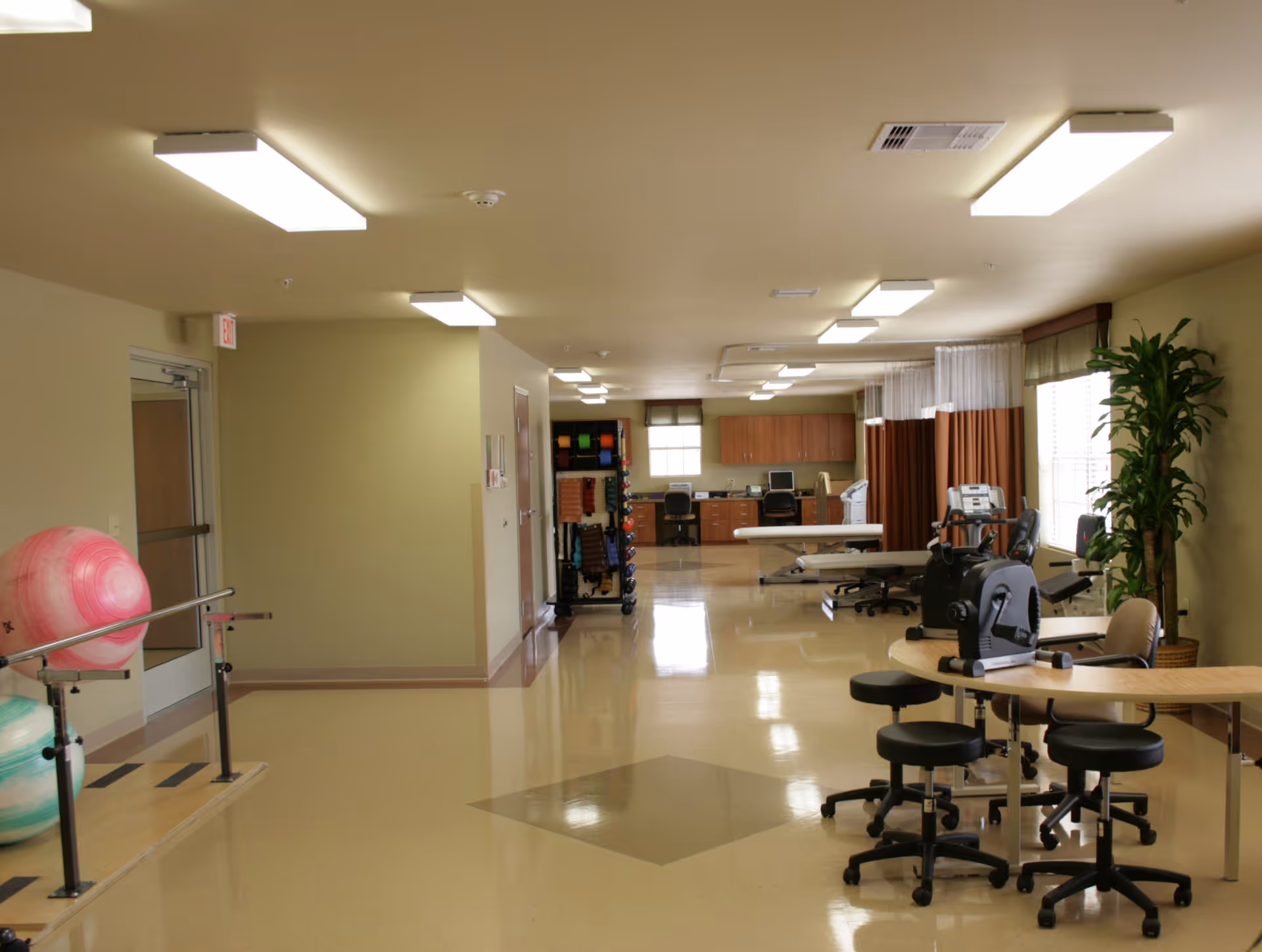 A spacious rehabilitation or physical therapy room with exercise equipment including large therapy balls, parallel bars, stationary exercise bikes, and treatment tables. The room has bright overhead lighting, beige walls, and a shiny floor. There are windows with curtains and a potted plant near the window.